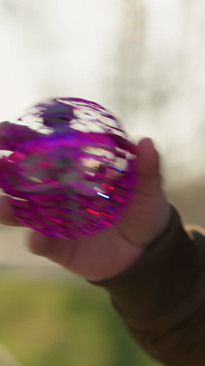 un primer plano de un adulto sosteniendo un juguete púrpura con un ventilador giratorio en el interior, el fondo muestra una vista borrosa de árboles y una pasarela