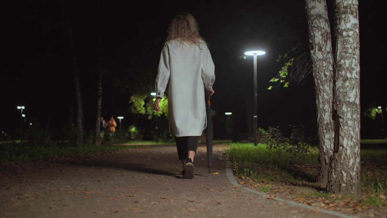 Back view of young girl holding umbrella walking along calm park path at night illuminated by soft streetlights, with two blurred figures walking in distant background among trees