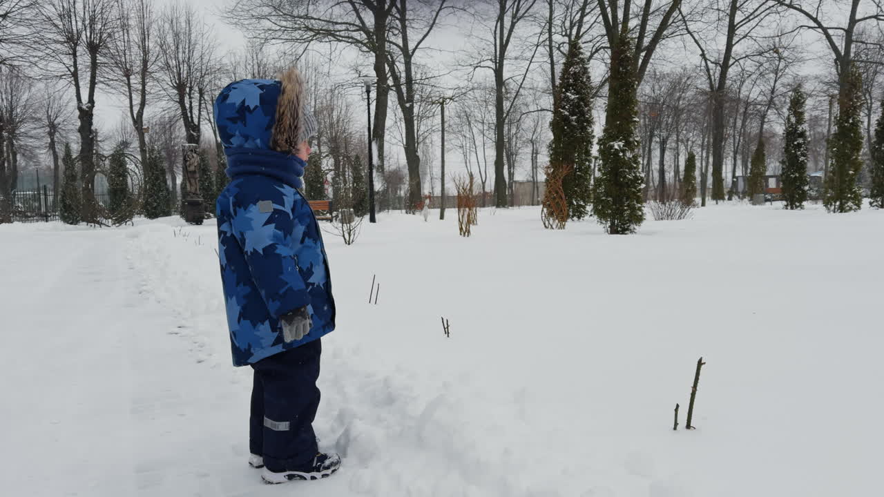 Caucasian toddler in blue clothes stands in winter. Side view. Baby boy points at something in front of him.