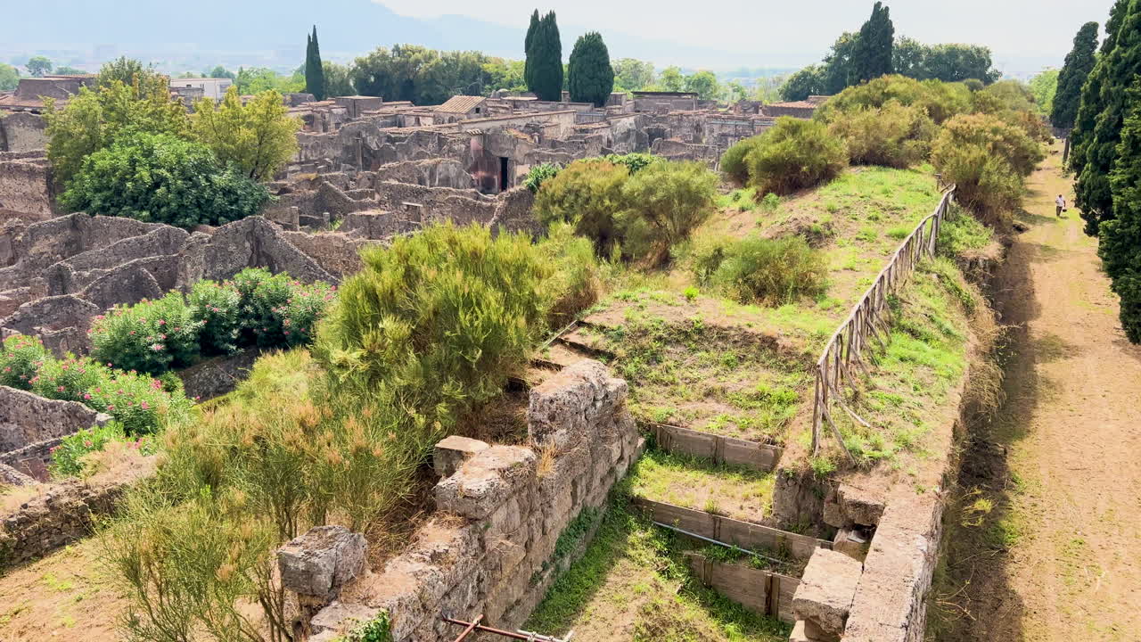 Panoramic view of the ruins of Pompeii with the city's ancient structures spread out under a clear sky, and the lush greenery contrasting with the stone remains