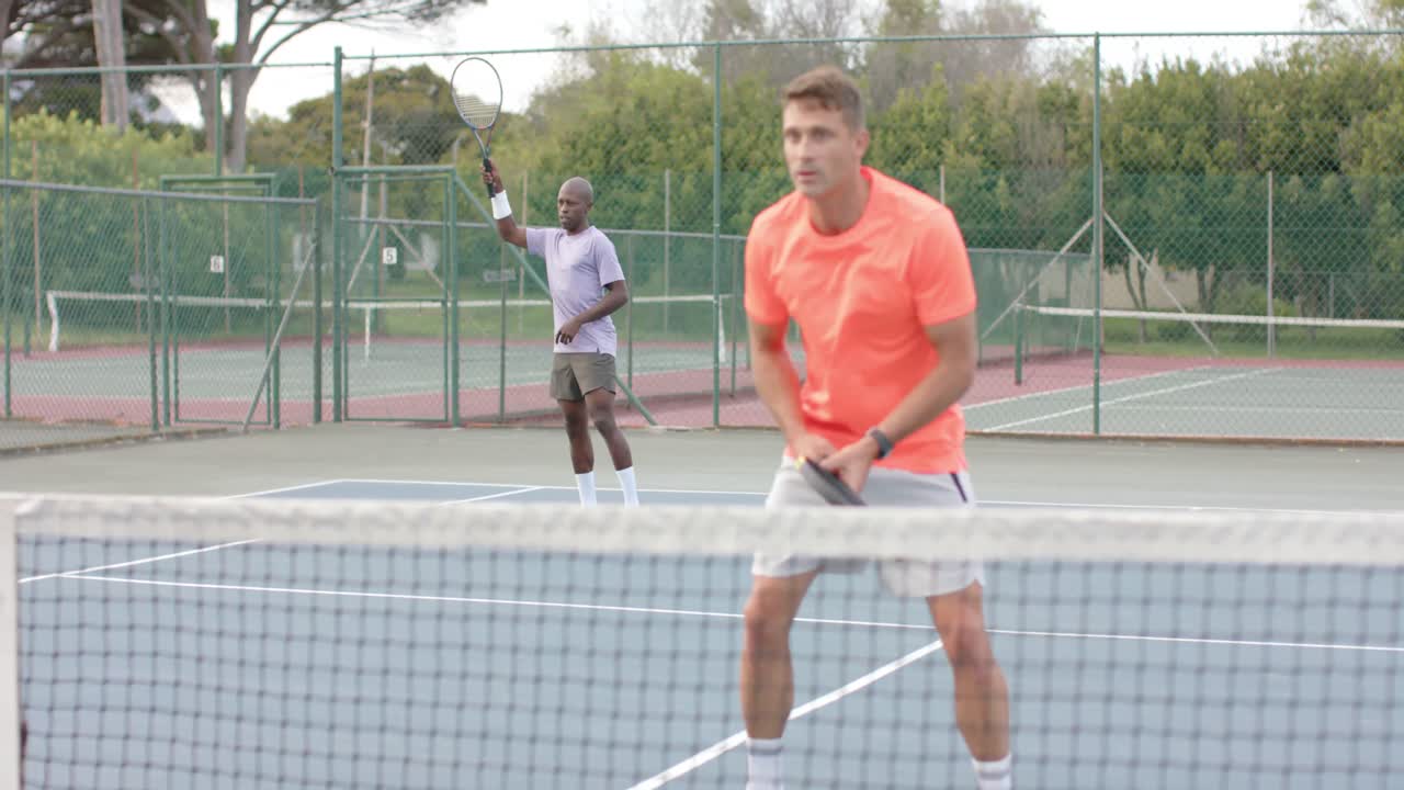 두 명의 다양한 남성 친구가 더블을 치며 느린 움직임으로 야외 코트에서 공을 네트 위로 돌려줍니다.