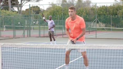 Two-diverse-male-friends-playing-doubles-returning-ball-over-net-at-outdoor-court-in-slow-motion