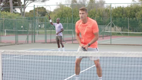 two diverse male friends playing doubles returning ball over net at outdoor court in slow motion