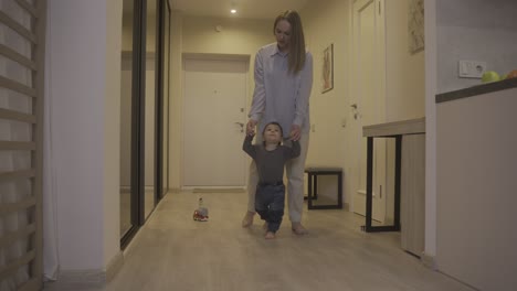 mother helping her baby to take his first steps from the corridor to the living room at home