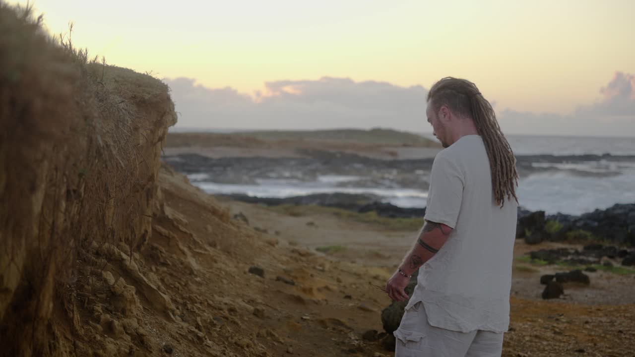 A man dressed in white stands still in profile on a rocky coastal path, calmly gazing toward the distant ocean horizon as waves break in the soft sunrise glow