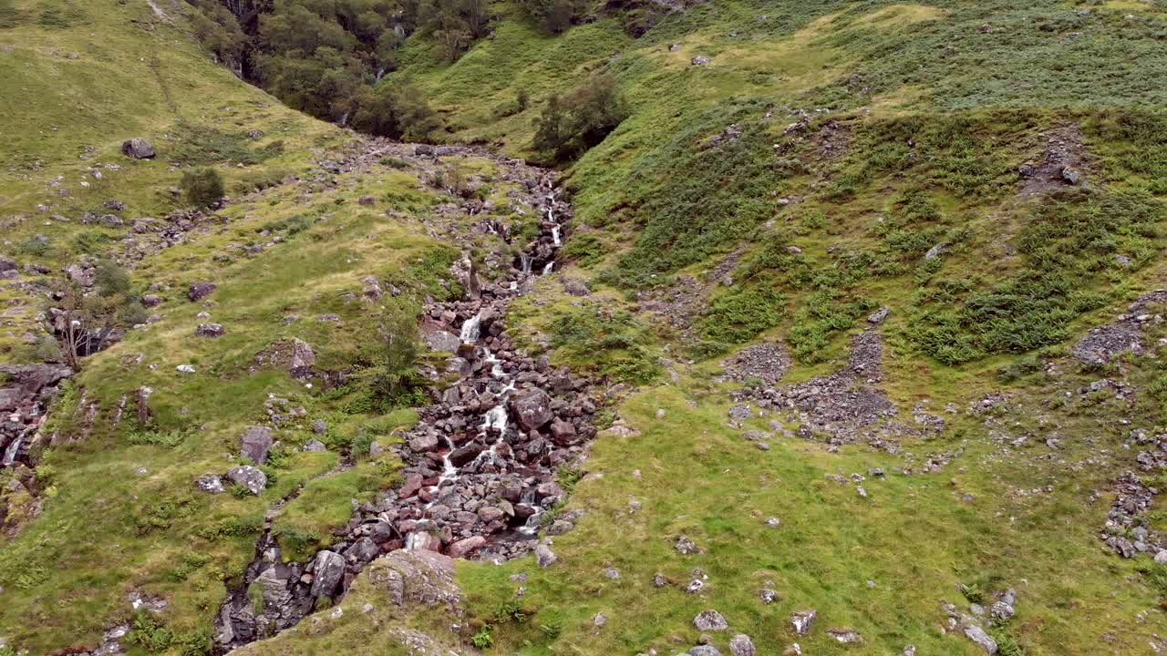 Vista aerea, che si allontana lentamente da un primo piano di un ripido ruscello roccioso sul versante lussureggiante e verdeggiante della valle di Glen Coe, Scozia.