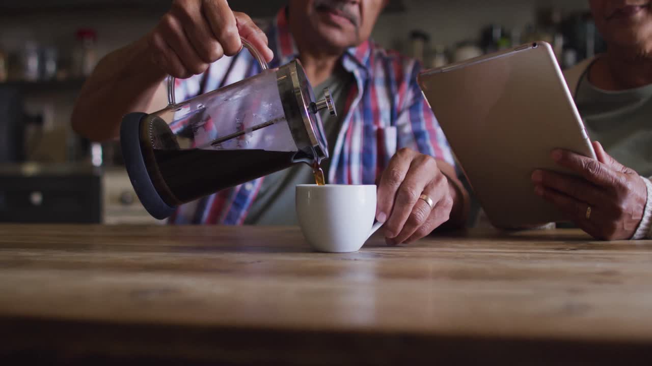 Pareja mayor mestiza sirviendo café y usando una tableta en la cocina