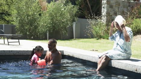 Un-Hombre-Birracial-Y-Un-Niño-Disfrutan-De-La-Piscina,-Una-Mujer-Con-Sombrero-Se-Sienta-A-Su-Lado