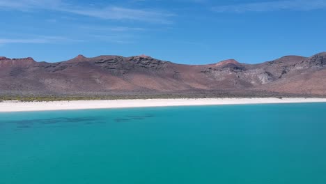 aerial video flying over beautiful tropical teal water toward a white sand beach at the foot of some small mountains