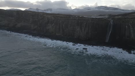 Kilt-rock-and-mealt-falls-on-isle-of-skye,-scotland-with-ocean-waves-and-snow-capped-mountains-in-the-backdrop,-aerial-view