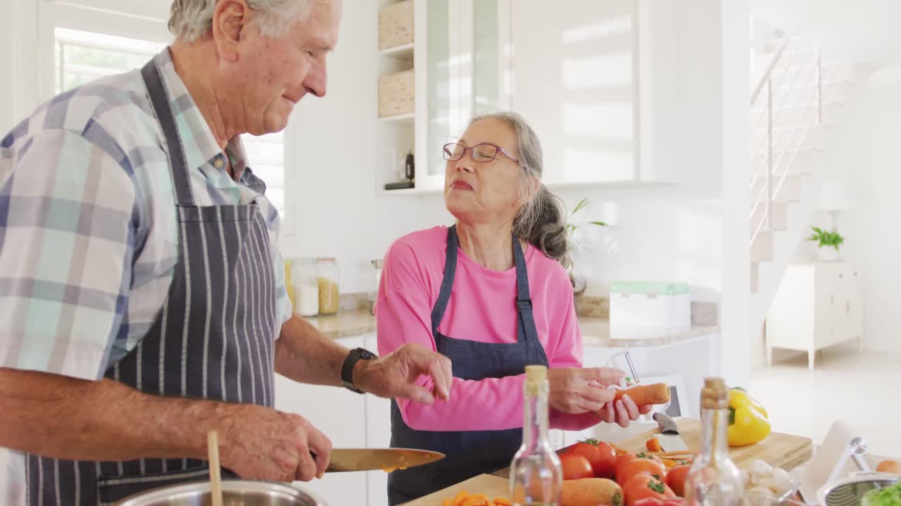 Felice coppia anziana multiculturale che indossa grembiuli e cucina in cucina.