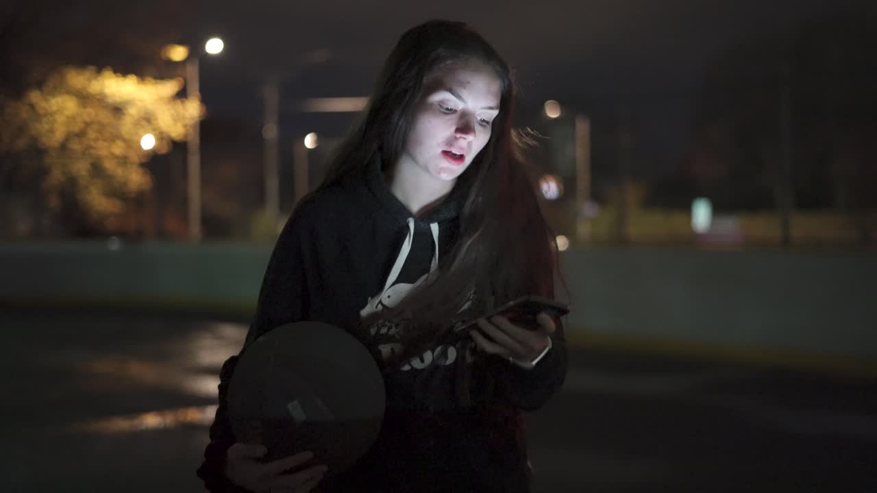 adolescente usando teléfono sosteniendo baloncesto en la cancha al aire libre por la noche en la ciudad