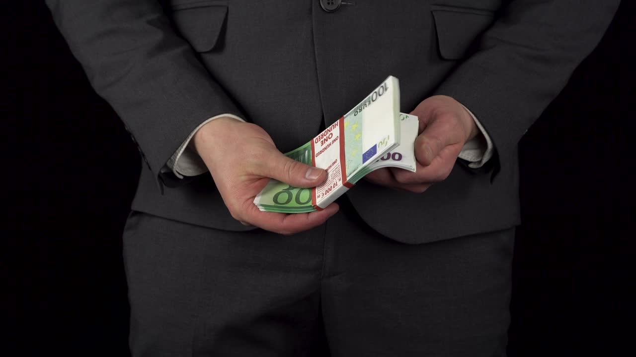 un joven empresario en traje saca un paquete de euros de su bolsillo y lo sostiene en sus manos. la cintura de un hombre de primer plano sobre un fondo negro.