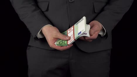 een jonge zakenman in een pak haalt een pakje euro's uit zijn zak en houdt het in zijn handen. de taille van een man close-up op een zwarte achtergrond.