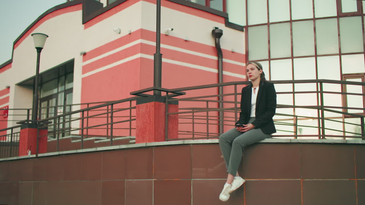 Woman in black suit waiting for someone seated on fence with phone in hand, legs crossed at ankle, in urban setting with modern glass building and lampposts