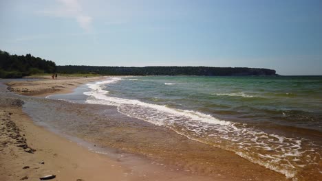 Sunny-beach-day-on-Gotland,-waves-gently-lapping-the-shore,-people-in-the-distance,-clear-blue-sky