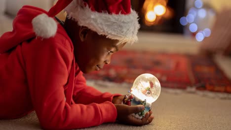 Animation-of-african-american-boy-in-santa-hat-holding-snow-globe-at-christmas-time