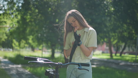 una mujer joven está de pie al lado de su bicicleta, poniéndose atentamente su guante de motociclista en un parque iluminado por el sol, la escena presenta un fondo verde pacífico y borroso con árboles y otro transeúnte