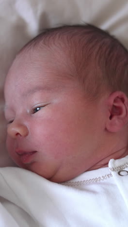 sweet newborn boy is lying on the bed and turning his little head back and forth. yawning tiny kid in a white costume. close-up from above. vertical video