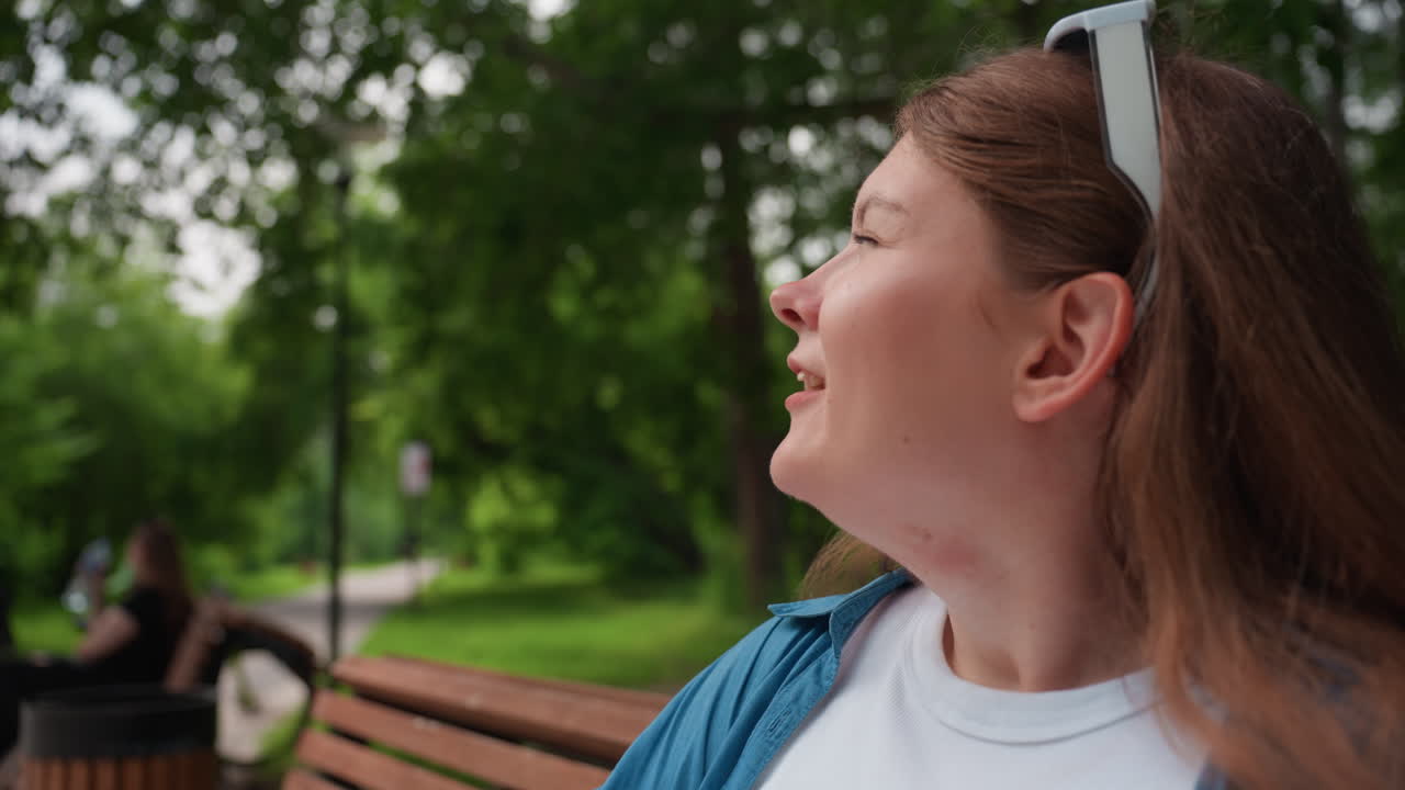 Close up of lady in casual outfit sitting outdoors on wooden bench sipping water from clear plastic bottle, relaxing under green trees on warm summer day