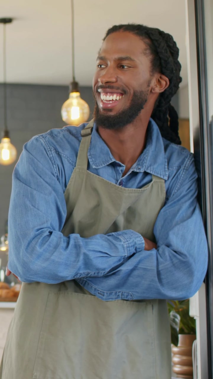 Video verticale: Barista sorridente appoggiato alla porta del bar, facendosi ombra, saluta dopo la chiamata di un cliente, con grembiule.