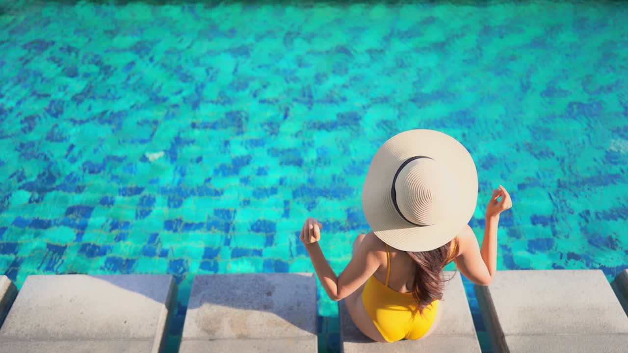 Backside view of the woman sitting on the edge of the swimming pool at an exotic hotel in Miami in yellow monokini and white hat