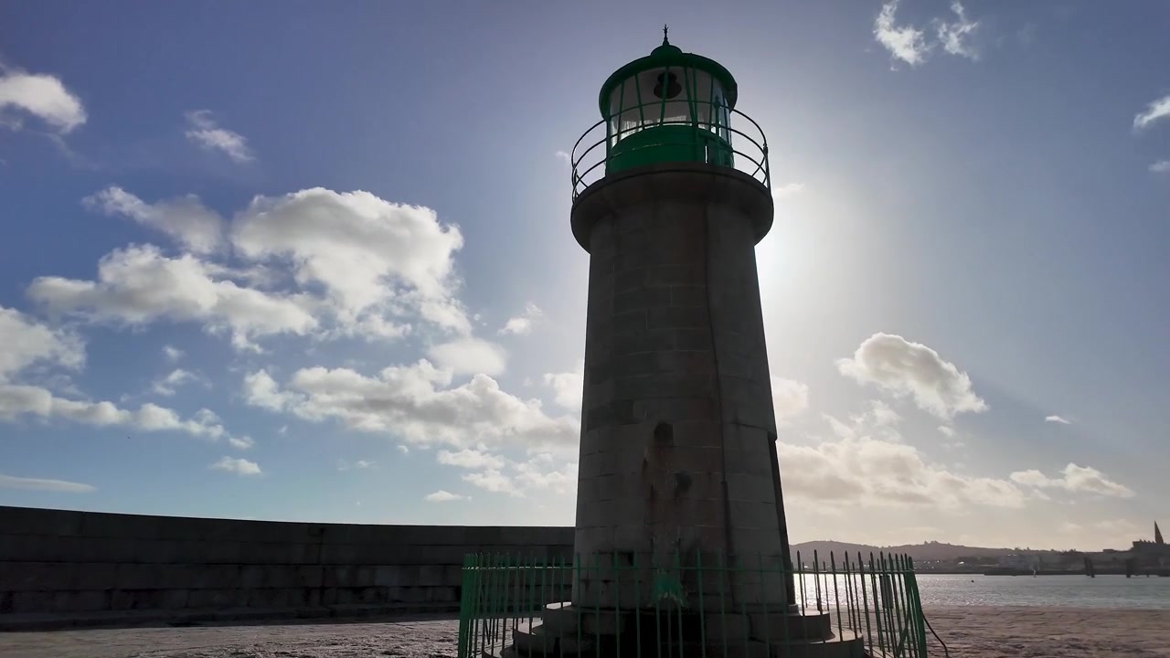 Avvicinamento a piedi all'iconico Faro di East Pier a Dún Laoghaire, Irlanda, con la luce del sole che risplende dietro la torre, paesaggio costiero e un'atmosfera marittima suggestiva.