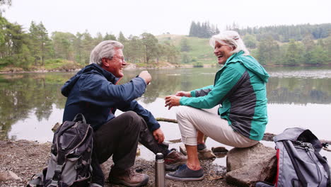 Pareja-Mayor-Sentada-Sobre-Las-Rocas-Junto-A-Un-Lago-Riendo-Y-Hablando-Durante-Unas-Vacaciones-En-Camping,-Lake-District,-Reino-Unido