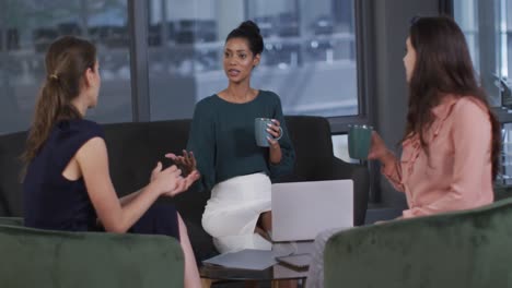 Diverse-group-of-three-businesswomen-drinking-coffee-and-talking-at-casual-office-meeting