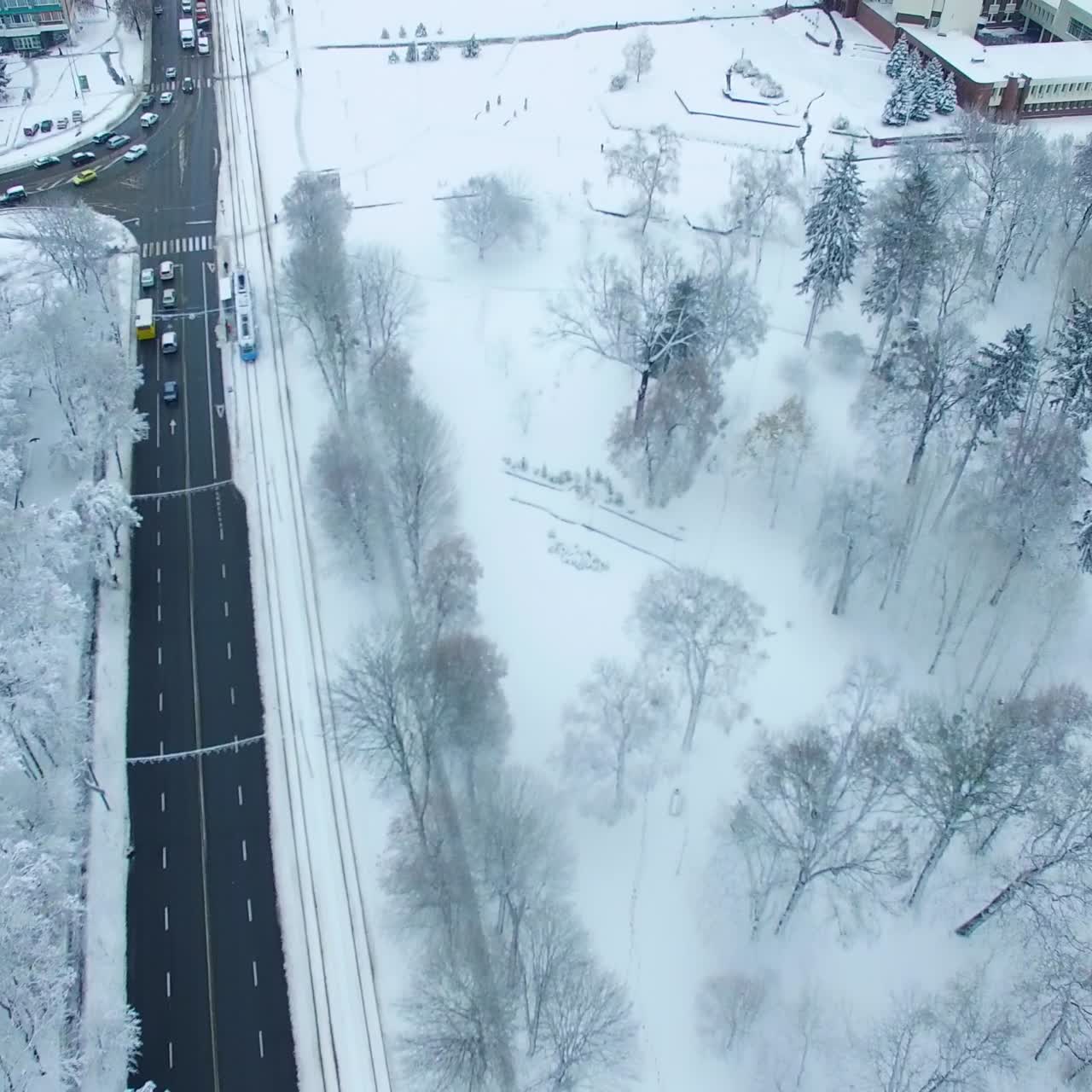 Flying over and along the road in the city covered with snow. Park with bare trees in snow. High buildings at backdrop