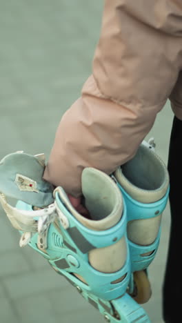 close-up shot of a girl dressed in a peach jacket and black trousers, carrying a pair of rollerblades as she walks along a paved park path during sunset.with the girl's face not visible