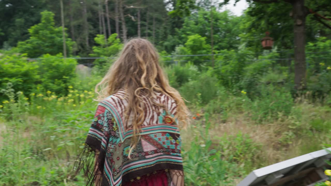 Giovane hippie che cammina di spalle alla telecamera attraverso un campo di fiori selvatici nella natura, stile di vita di ecovillaggio e tiny house con agricoltura biologica sostenibile.