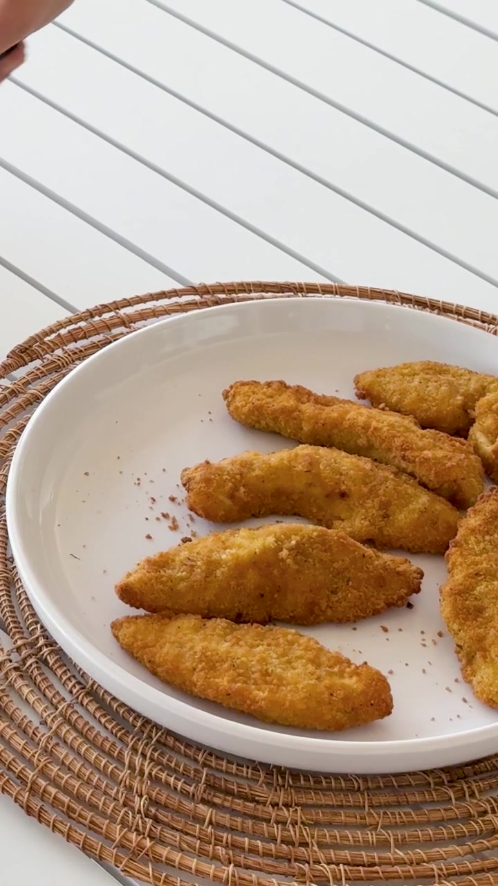 A hand uses black tongs to pick up golden breaded chicken tenders from a plate.