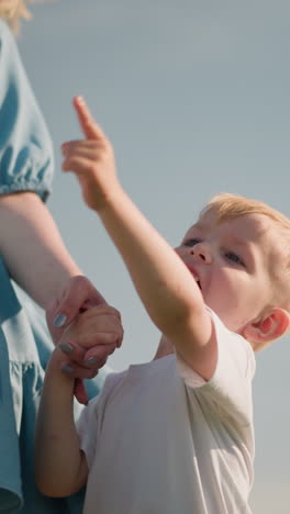a little boy in a white shirt smiles pointing at the environment, holding his mother s hand, who is wearing a blue dress. another boy is partially visible in the background