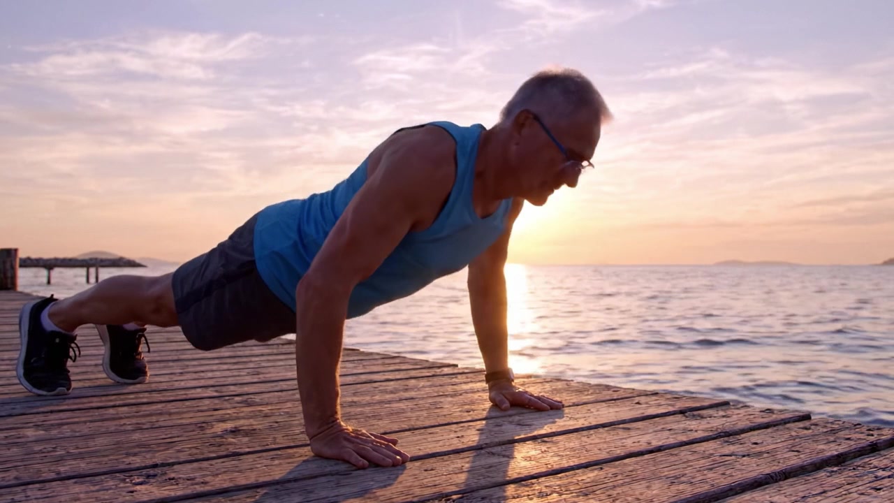 A focused older man performing push-ups on a wooden dock at sunset, showcasing determination and fitness against a picturesque background of a tranquil sea.
