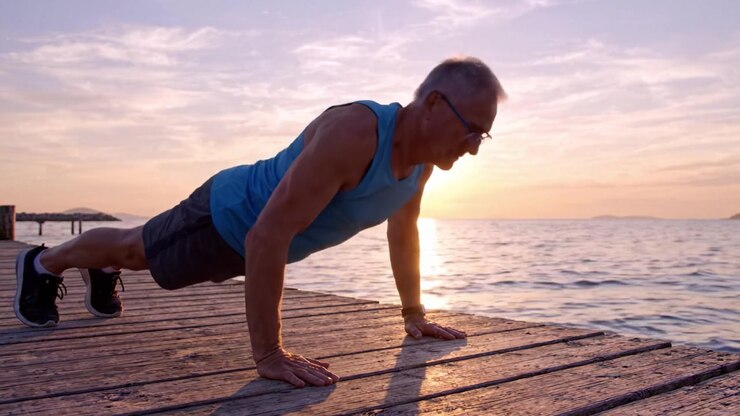 A focused older man performing push-ups on a wooden dock at sunset, showcasing determination and fitness against a picturesque background of a tranquil sea.