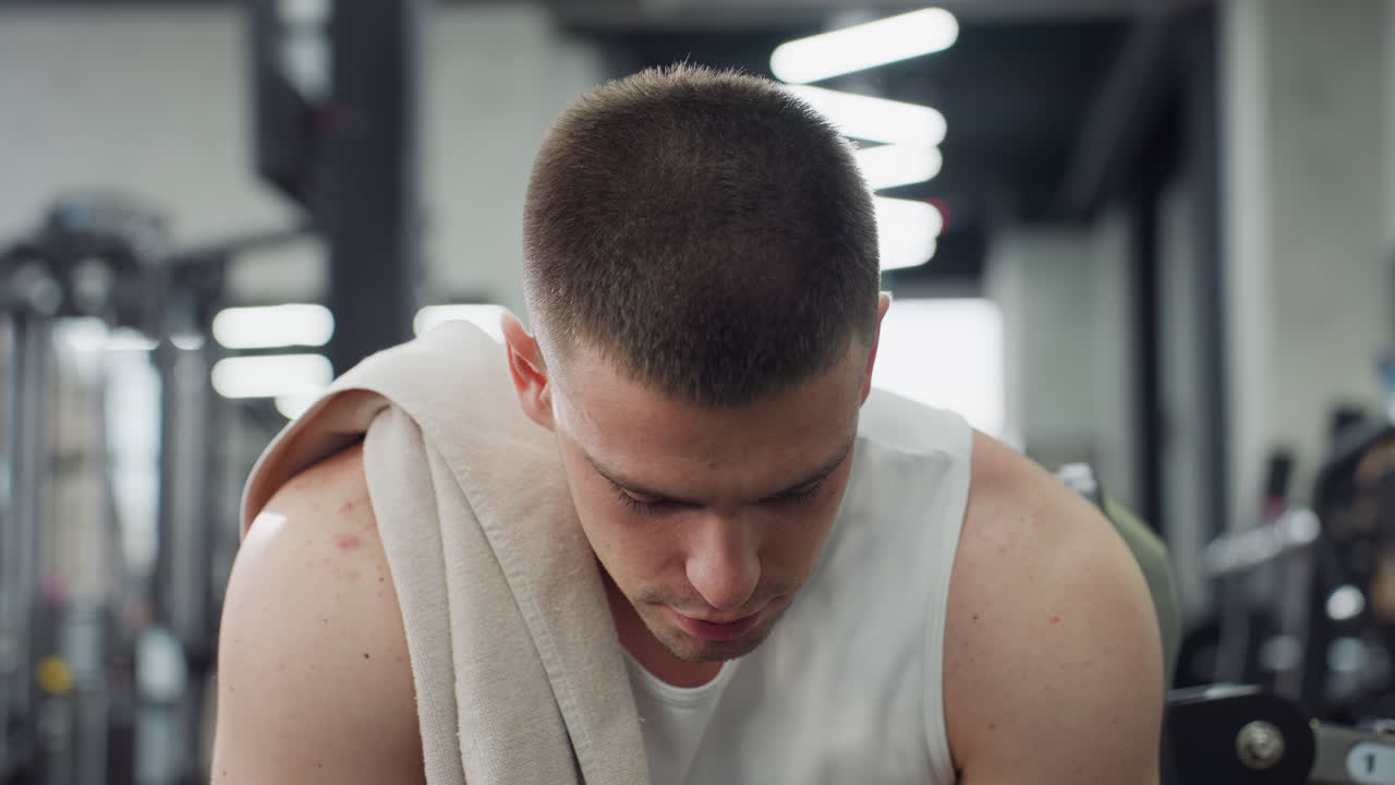 Portrait of fitness enthusiast sits on weight bench with towel draped over shoulder exhaling relief after intense chest press workout under modern gym lights showing focused calm expression