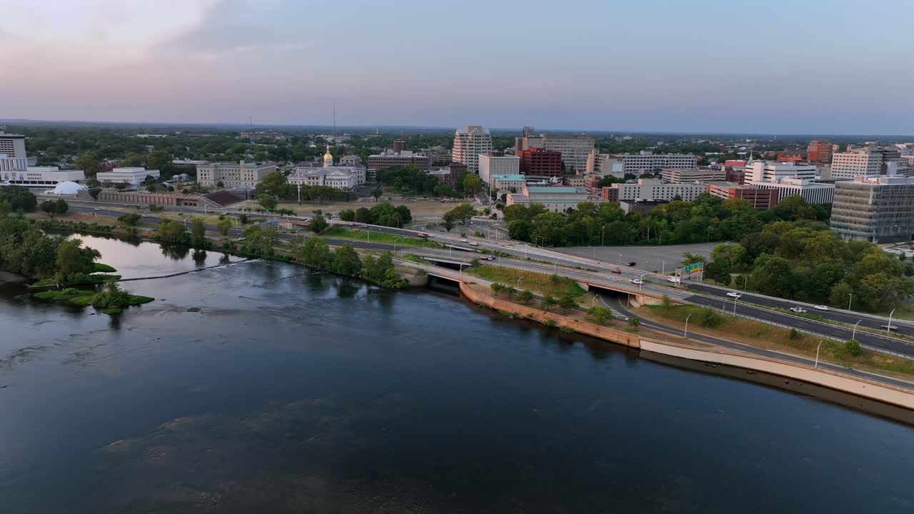 Aerial flyover river in Trenton city at sunset time. Tranquil waters and traffic on shoreline road. Downtown with towers and buildings in background. Wide shot. New Jersey in summer season