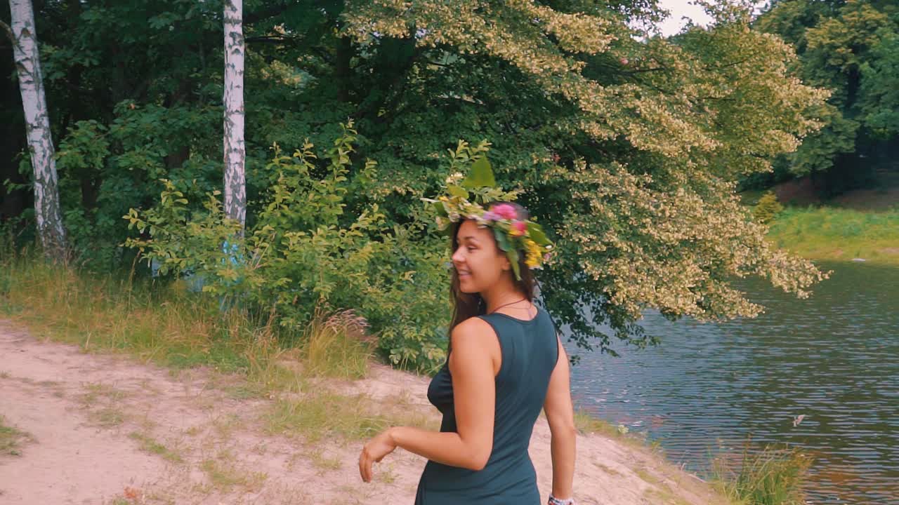 cámara lenta de mujer joven sensual feliz con corona de flores por el lago y el árbol