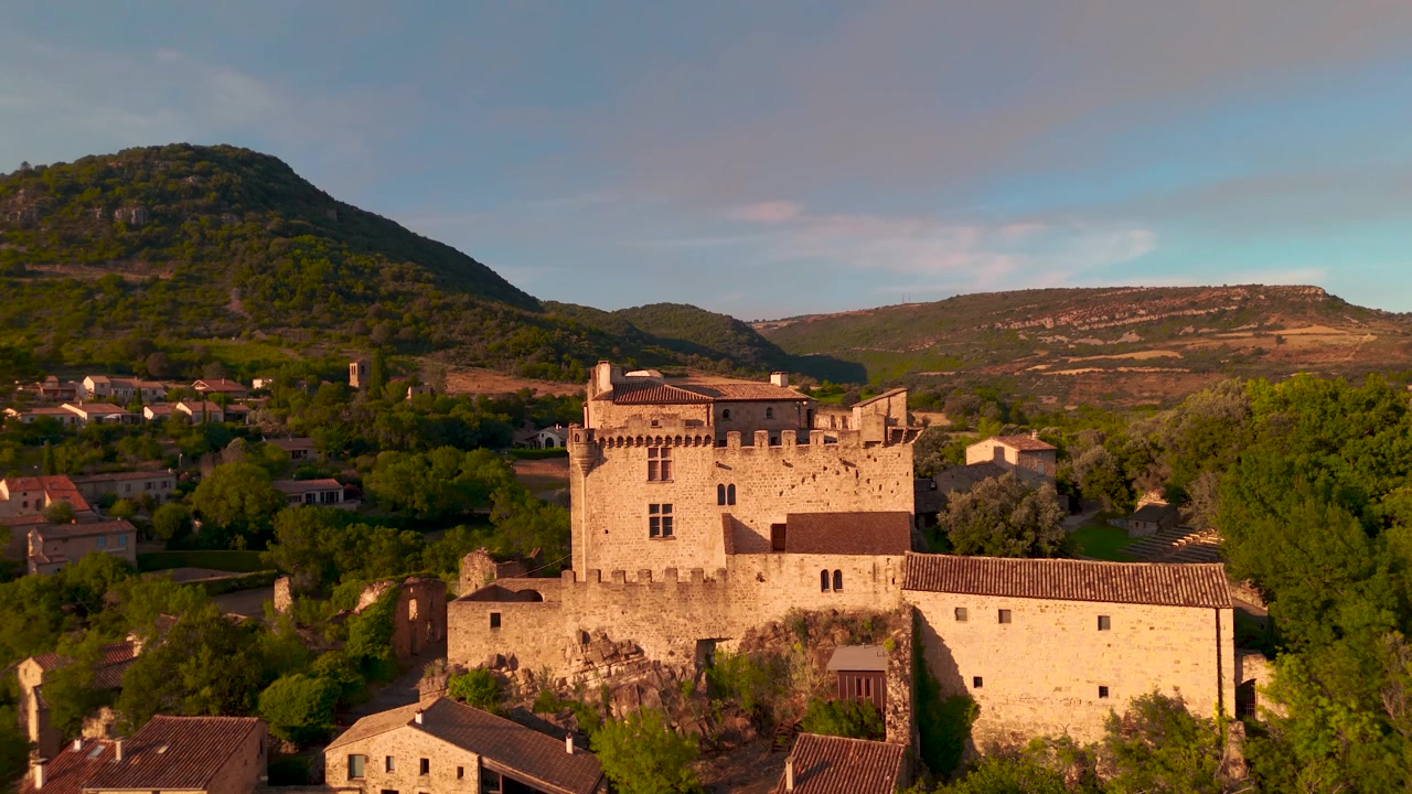 Castello e villaggio storico circondati da colline boscose nella luce dorata della sera, ripresa aerea in avvicinamento.