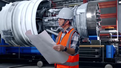 side view of asian male engineer with safety helmet looking at blueprint in his hands and looking around while standing with airplane engine maintenance conducted, analytics checking the turbine