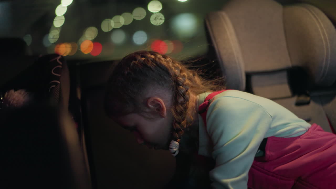 A close view of a child turning on the light in a car to search for something, with a background of blurred car lights visible through the window