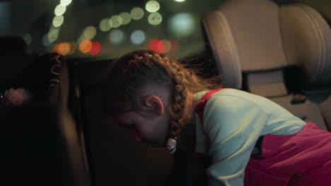 a close view of a child turning on the light in a car to search for something, with a background of blurred car lights visible through the window