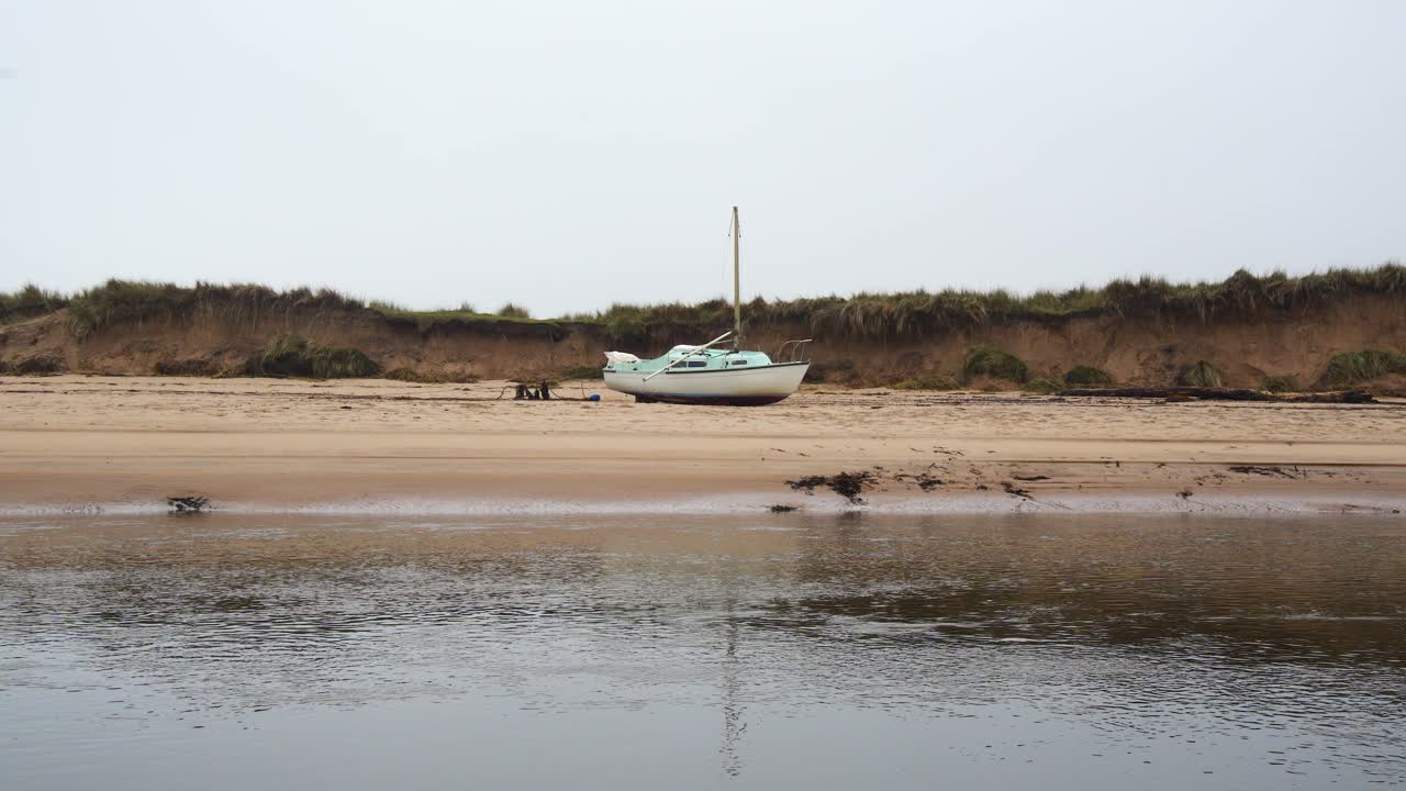 A sailboat rests on a deserted beach near calm waters under an overcast sky