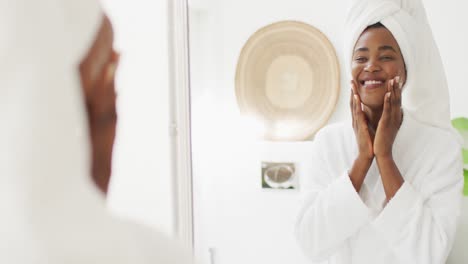 Video-of-happy-african-american-woman-in-robe-looking-at-mirror-and-moisturizing-face