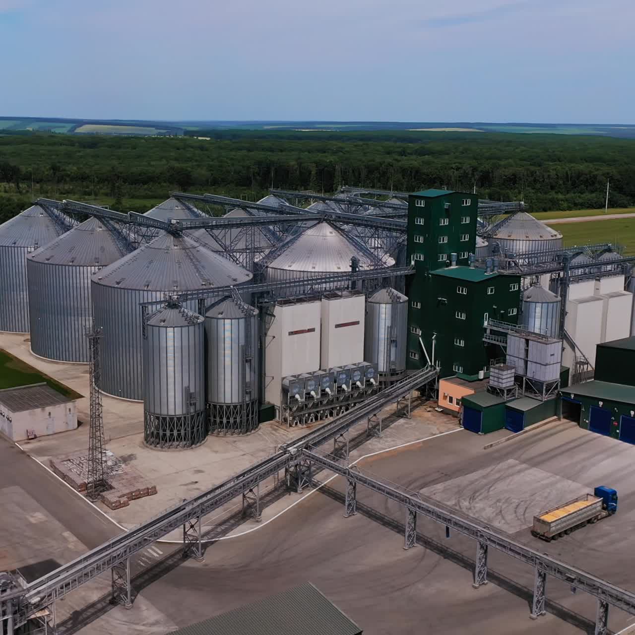 Commercial grain silos outdoors. Flight from the industrial building of the plant to the large metal tanks of elevator. Aerial view. Woods and farmlands at the backdrop