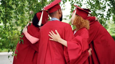 joyful friends graduating students in gowns and mortarboards are congratulating each other after graduation ceremony, they are hugging and laughing.