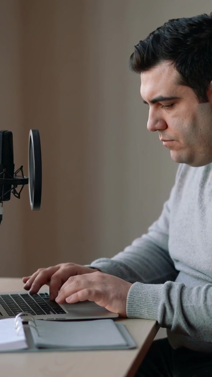Video vertical: Enfoque en un portátil, hombre con suéter escribiendo en el escritorio de casa, con micrófono y filtro antipop visibles.