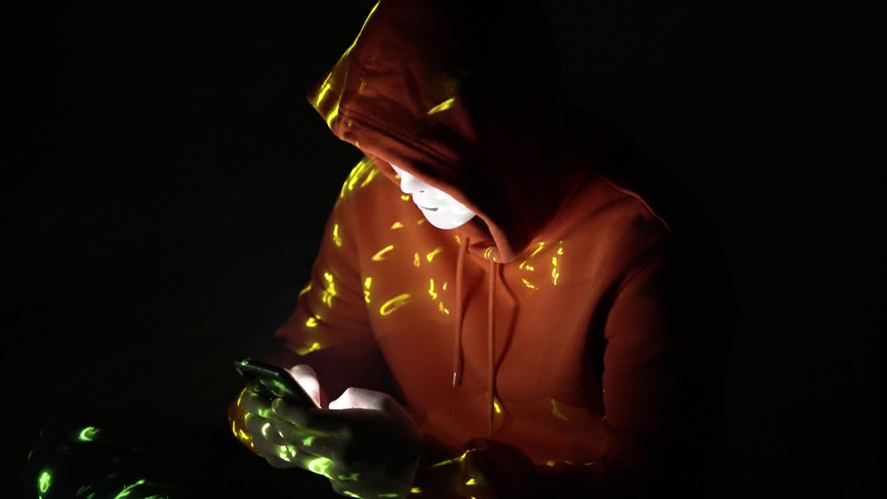 In a pitch-black room, a hooded figure in a white mask stares at a smartphone, illuminated by scattered yellow digital fragments, evoking cyber secrecy and mobile-based intrusion attempts