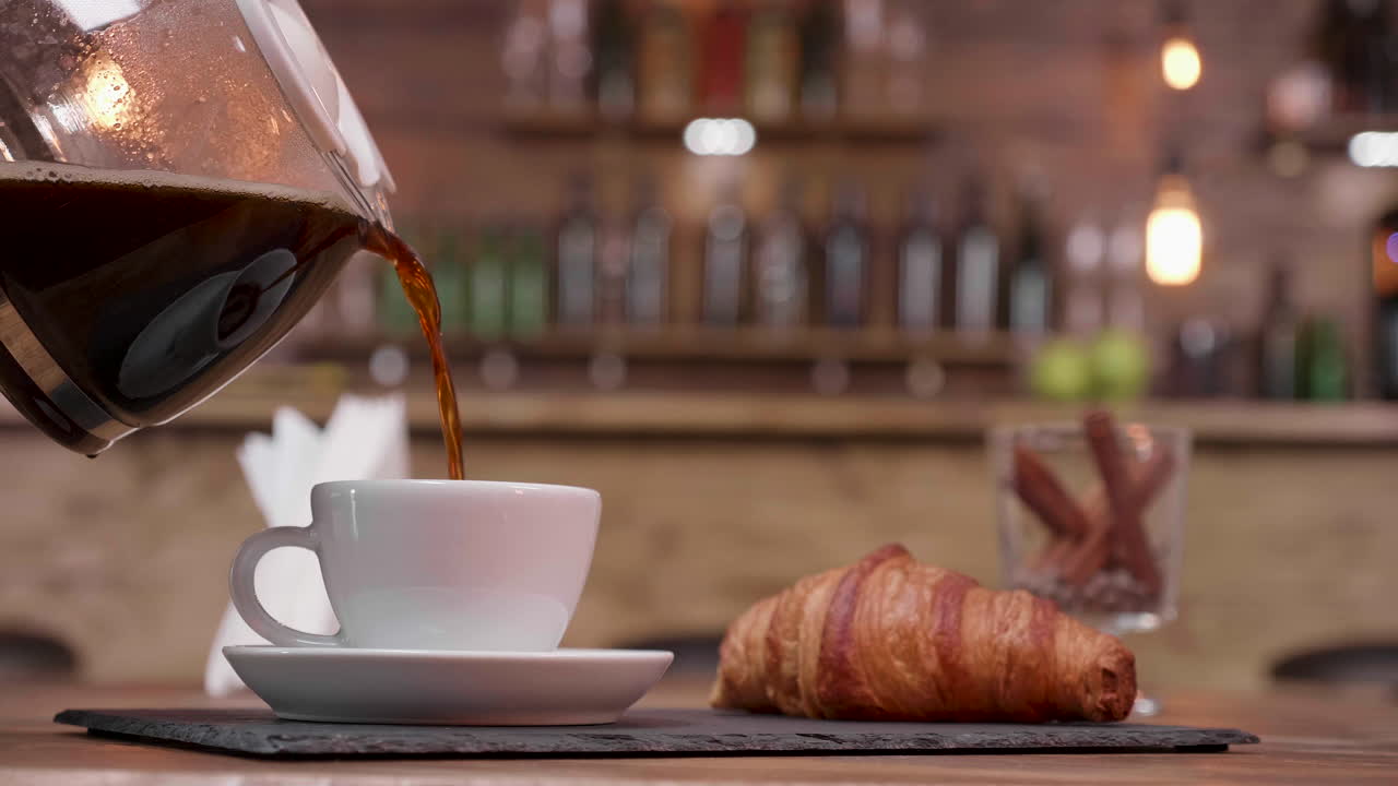 Coffee being poured into a cup with a croissant on the side